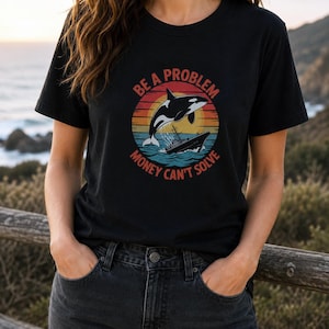 Könnte beinhalten: Ein schwarzes Kurzarm-T-Shirt mit einem Grafikdruck eines Orcas, der über ein Boot springt, vor einem Sonnenuntergangshintergrund. Der Text "BE A PROBLEM" ist darüber und "MONEY CAN'T SOLVE" darunter in einer fetten Schriftart gebogen.