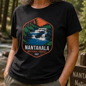 May include: Black t-shirt with a hexagonal graphic depicting a waterfall scene. The design includes the text "Nantahala National Forest - 1907". The shirt is made of a soft material, suitable for outdoor activities. The design is in shades of blue, green, and orange.