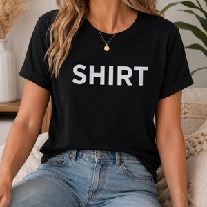 Puede incluir: Camiseta negra con la palabra "SHIRT" impresa en letras blancas. La camiseta tiene mangas cortas y cuello redondo. La modelo lleva vaqueros azules y un collar dorado.
