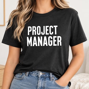 Puede incluir: Una camiseta negra de manga corta con el texto PROJECT MANAGER impreso en letras mayúsculas blancas y negritas en el pecho. La camiseta tiene un cuello redondo clásico y se combina con unos vaqueros azules con un botón metálico visible.