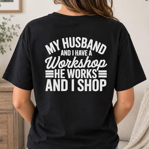 以下が含まれることがあります： 背面に白い文字で「MY HUSBAND AND I HAVE A Workshop HE WORKS AND I SHOP」とプリントされた黒い半袖Tシャツ。クルーネック仕様で、背面から撮影されており、グラフィックプリントがはっきりと確認できるデザインとなっている。