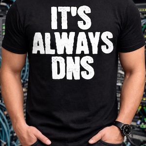 May include: A black short-sleeved t-shirt featuring the white distressed-style text "IT'S ALWAYS DNS" printed in a bold, sans-serif font across the chest. The garment has a classic crew neckline and a straight hemline, providing a standard fit.