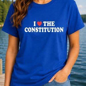 Puede incluir: Una camiseta de manga corta de color azul real con el texto blanco "I" seguido de un símbolo de corazón rojo y el texto blanco "THE CONSTITUTION" impreso en el pecho. La camiseta tiene cuello redondo y un corte estándar de tela lisa.
