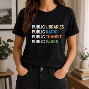 May include: Black t-shirt with the words "PUBLIC LIBRARIES, PUBLIC RADIO, PUBLIC TRANSIT, PUBLIC PARKS" in a stacked format. The text is in a mix of colors: beige, blue, orange, and green. The person is wearing black jeans.