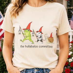 Op de afbeelding: Een crèmekleurig t-shirt met een afbeelding van een groene kikker, een opossum en een slak, die allemaal rode puntmutsen dragen. De kikker en de opossum dragen lantaarns aan stokken. Onder de illustratie staat de tekst, the hullabaloo committee.