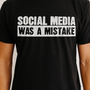 Puede incluir: Camiseta negra con el texto blanco "SOCIAL MEDIA WAS A MISTAKE" en una fuente en negrita sin serifa. El texto está centrado en el pecho. La camiseta tiene mangas cortas y cuello redondo.