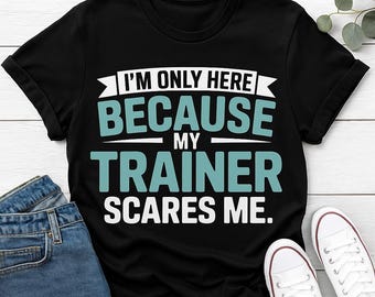 Peut inclure: T-shirt noir avec l'inscription "I'M ONLY HERE BECAUSE MY TRAINER SCARES ME" en blanc et turquoise. L'image comprend un jean bleu et des baskets blanches, suggérant un thème lié au fitness ou à la salle de sport.