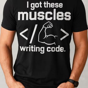 Könnte beinhalten: Schwarzes T-Shirt mit weißem Text: „Ich habe diese Muskeln beim Schreiben von Code“. Das Design beinhaltet eine Grafik eines Bizeps und Codesymbole. Das T-Shirt wird von einer Person in blauen Jeans getragen.