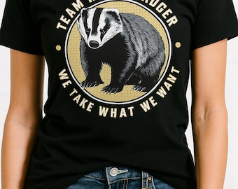 May include: Black t-shirt featuring a honey badger illustration inside a gold circle. The text around the circle reads "TEAM HONEY BADGER" and "WE TAKE WHAT WE WANT". The shirt is worn with blue jeans.