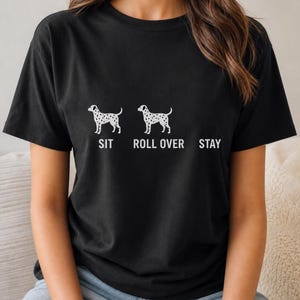 以下が含まれることがあります： 黒の半袖Tシャツで、2匹の斑点のある犬の白いグラフィックが描かれています。最初の犬の下には「SIT」、2匹目の犬の下には「ROLL OVER」、2匹目の犬の右側には「STAY」という白い文字がプリントされています。