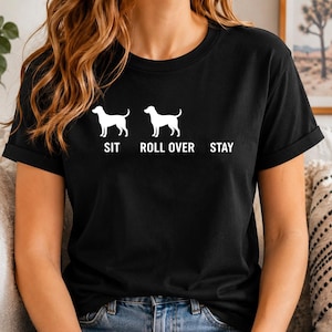 以下が含まれることがあります： 黒の半袖Tシャツで、2匹の犬のシルエットが白でプリントされています。犬の下には「SIT」と「ROLL OVER」という文字があり、右側には犬のシルエットがない状態で「STAY」という文字が配置されています。