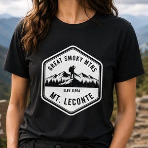 May include: A black short-sleeved t-shirt featuring a white hexagonal graphic print. The design depicts a hiker on a mountain peak with the text GREAT SMOKY MTNS, ELEV. 6,594, and MT. LECONTE. The graphic has a distressed, weathered texture.