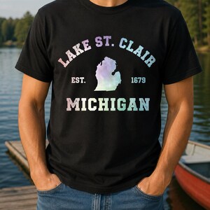 Puede incluir: Una camiseta negra de manga corta con un gráfico de estilo acuarela en tonos pastel del estado de Michigan. El texto dice LAKE ST. CLAIR, EST. 1679 y MICHIGAN en una fuente desgastada, a juego con la estética colorida y difuminada.