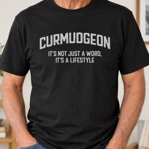 以下が含まれることがあります： 胸元に大きなかすれた白文字でCURMUDGEONとプリントされ、その下に小さくIT'S NOT JUST A WORD, IT'S A LIFESTYLEと書かれた黒の半袖Tシャツ。クルーネックで標準的なフィット感のTシャツです。
