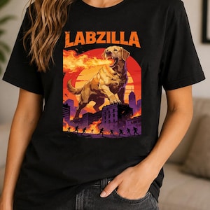 May include: A black t-shirt featuring a graphic print of a giant golden Labrador dog breathing fire over a city skyline. The text LABZILLA is printed in orange above the dog, with silhouettes of people running below against a large red sun.