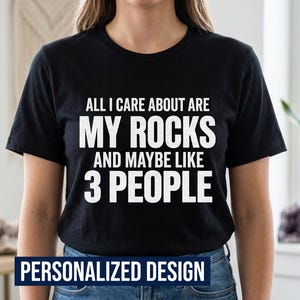 以下が含まれることがあります： ALL I CARE ABOUT ARE MY ROCKS AND MAYBE LIKE 3 PEOPLEという白い文字がプリントされた黒い半袖Tシャツ。クルーネックの標準的なフィット感で、胸元のグラフィックデザインがはっきりと確認できるデザインとなっています。
