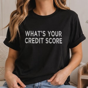 May include: Black t-shirt featuring the text "WHAT'S YOUR CREDIT SCORE" in a white, distressed font. The shirt has short sleeves and a relaxed fit. The style is casual and comfortable, suitable for everyday wear.