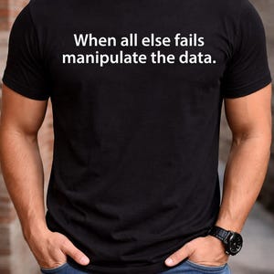 May include: Black t-shirt with the white text "When all else fails manipulate the data." The shirt is paired with blue jeans. A black watch is visible on the left wrist. The person has their hands in their pockets.