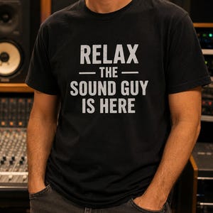 Könnte beinhalten: Ein schwarzes Kurzarm-T-Shirt mit weißem Text im Used-Look auf der Brust, der RELAX — THE — SOUND GUY IS HERE lautet. Das Shirt hat einen Rundhalsausschnitt, eine normale Passform und einen geraden Saum aus einfarbigem schwarzem Stoff.