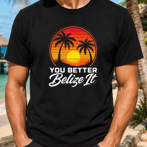 May include: A black short-sleeved t-shirt featuring a circular graphic of a sunset with two palm trees in silhouette. Below the graphic, the text reads "YOU BETTER Belize It" in a bold, white, stylized font centered on the chest area.