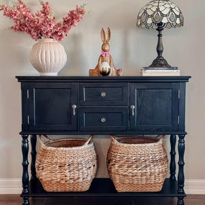 May include: A black wooden sideboard with two doors, two drawers, and two woven baskets. The top is decorated with a pink vase of flowers, a wooden bunny holding a golden egg, and a stained glass lamp.