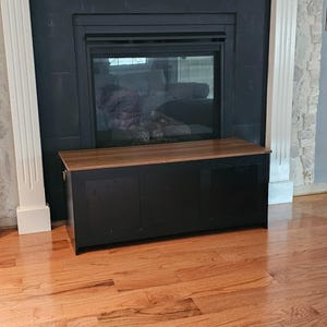 Puede incluir: Un arc&oacute;n de almacenamiento negro con una tapa de madera se encuentra frente a una chimenea. El arc&oacute;n tiene tres paneles y forma rectangular. La chimenea es negra con frente de vidrio y el suelo es de madera noble.