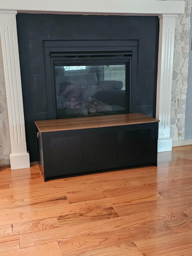 Puede incluir: Un arc&oacute;n de almacenamiento negro con una tapa de madera se encuentra frente a una chimenea. El arc&oacute;n tiene tres paneles y forma rectangular. La chimenea es negra con frente de vidrio y el suelo es de madera noble.