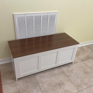 Puede incluir: Un arc&oacute;n de almacenamiento de madera blanca con una tapa marr&oacute;n oscuro. El arc&oacute;n tiene tres paneles rectangulares en la parte delantera y est&aacute; colocado frente a una rejilla de ventilaci&oacute;n blanca. El arc&oacute;n est&aacute; sobre un suelo embaldosado.