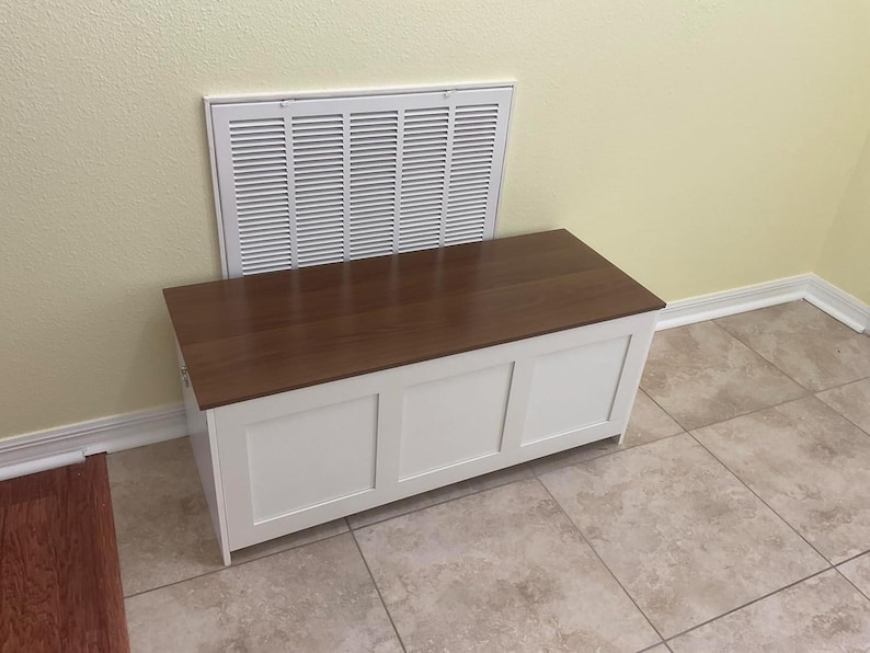 Puede incluir: Un arc&oacute;n de almacenamiento de madera blanca con una tapa marr&oacute;n oscuro. El arc&oacute;n tiene tres paneles rectangulares en la parte delantera y est&aacute; colocado frente a una rejilla de ventilaci&oacute;n blanca. El arc&oacute;n est&aacute; sobre un suelo embaldosado.