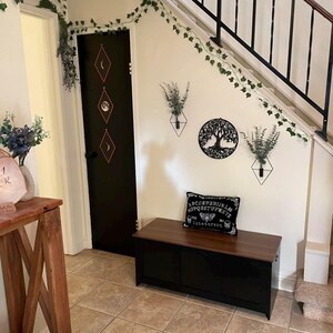 Puede incluir: Interior de una casa con un banco de almacenamiento negro con tapa de madera, una puerta negra con dise&ntilde;os de diamantes y una decoraci&oacute;n de pared con un &aacute;rbol de la vida. Enredaderas y plantas verdes adornan las paredes y el marco de la puerta.