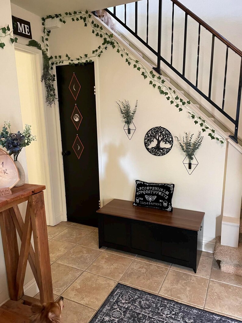 Puede incluir: Interior de una casa con un banco de almacenamiento negro con tapa de madera, una puerta negra con dise&ntilde;os de diamantes y una decoraci&oacute;n de pared con un &aacute;rbol de la vida. Enredaderas y plantas verdes adornan las paredes y el marco de la puerta.