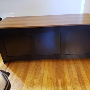 Puede incluir: Un arc&oacute;n de almacenamiento rectangular de madera con una base marr&oacute;n oscuro y una parte superior marr&oacute;n claro. El arc&oacute;n tiene un dise&ntilde;o frontal con paneles y est&aacute; colocado sobre un suelo de madera. El arc&oacute;n es un mueble.