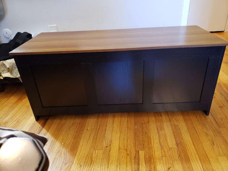 Puede incluir: Un arc&oacute;n de almacenamiento rectangular de madera con una base marr&oacute;n oscuro y una parte superior marr&oacute;n claro. El arc&oacute;n tiene un dise&ntilde;o frontal con paneles y est&aacute; colocado sobre un suelo de madera. El arc&oacute;n es un mueble.