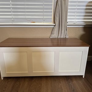Puede incluir: Un arc&oacute;n rectangular con tapa de madera marr&oacute;n oscuro y frente de paneles blancos. El arc&oacute;n est&aacute; colocado frente a una ventana con persianas. El arc&oacute;n mide aproximadamente 122 cm de ancho.
