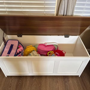 Puede incluir: Un arc&oacute;n de almacenamiento de madera blanca con tapa marr&oacute;n oscuro, parcialmente abierto. El arc&oacute;n est&aacute; lleno de varios juguetes para ni&ntilde;os, incluyendo una bolsa rosa, una bolsa amarilla y un peluche rojo.