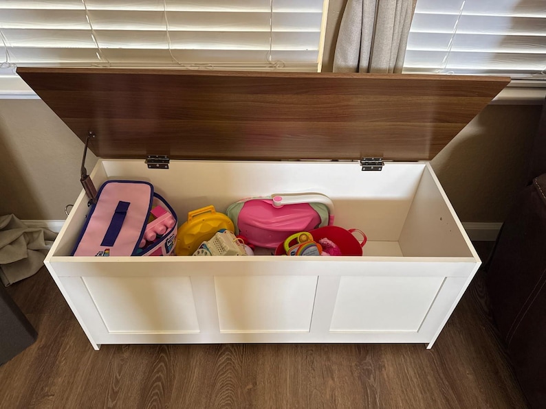 Puede incluir: Un arc&oacute;n de almacenamiento de madera blanca con tapa marr&oacute;n oscuro, parcialmente abierto. El arc&oacute;n est&aacute; lleno de varios juguetes para ni&ntilde;os, incluyendo una bolsa rosa, una bolsa amarilla y un peluche rojo.