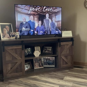 May include: A dark brown wooden TV stand with sliding barn doors. The TV displays the words "pure love" and a group of men in suits. Several framed photos and decorative items are on the stand.