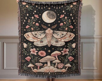 May include: A black woven throw blanket featuring a large central moth with beige and brown wings, a crescent moon above, and various mushrooms and pink flowers below. The blanket has a decorative border and fringed edges on the top and bottom.