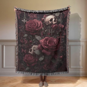 Könnte beinhalten: Eine dunkle Wandteppichdecke mit einem gotischen Design. Sie zeigt Schädel, Rosen und Kreuze auf schwarzem Hintergrund. Große burgunderfarbene Rosen und detaillierte Schädelmotive sind prominent. Die Decke hat einen Fransenrand.