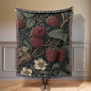 May include: A woven throw blanket featuring a botanical design of ripe red raspberries, green leaves, and white flowers against a dark green background. The blanket has a fringed edge and is held up by a person.