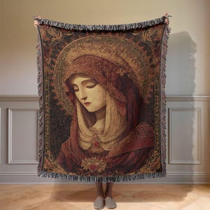 May include: A woven tapestry throw blanket depicting a portrait of a woman with a halo and a red cloak. The artwork has a vintage aesthetic with a detailed design and fringe edges. The colour palette includes reds, golds, and browns.