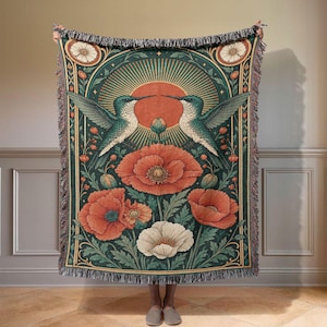 May include: A woven throw blanket featuring a symmetrical design of two hummingbirds facing each other above large orange and white poppy flowers. The background is a dark teal with a central orange sunburst motif and decorative floral borders.
