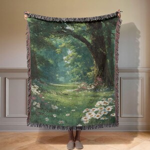 Op de afbeelding: Een geweven sprei met een serene bosscène met een pad dat in de verte leidt. De deken toont een weelderig groen landschap met bomen, wilde bloemen en een gefranjerde rand. Het kleurenpalet omvat groen, wit en bruin.