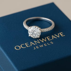 May include: A silver engagement ring with a large, round, clear gemstone. The band is encrusted with smaller stones. The ring rests on a blue box with the gold text "OCEANWEAVE JEWELS".