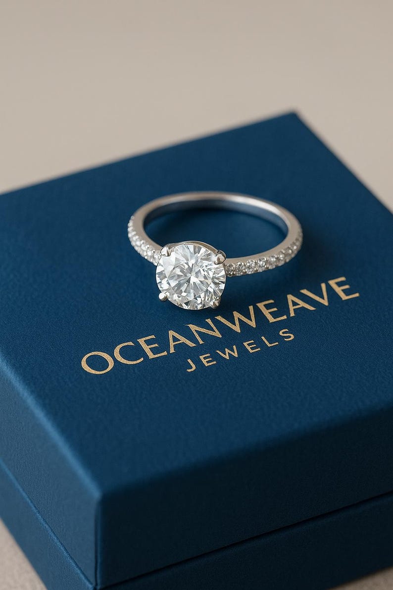 May include: A silver engagement ring with a large, round, clear gemstone. The band is encrusted with smaller stones. The ring rests on a blue box with the gold text "OCEANWEAVE JEWELS".