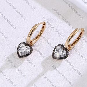 May include: A pair of gold hoop earrings with heart-shaped pendants. Each pendant features a clear, heart-cut stone set within a dark metal frame. The earrings are displayed on a white surface.