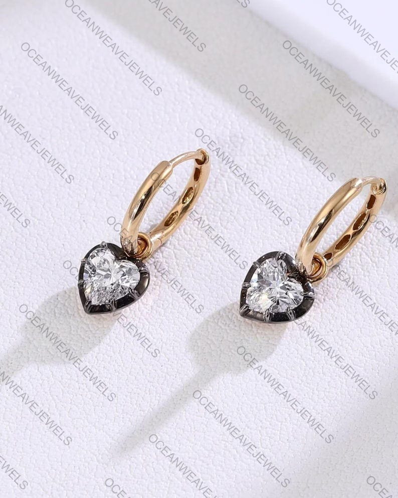 May include: A pair of gold hoop earrings with heart-shaped pendants. Each pendant features a clear, heart-cut stone set within a dark metal frame. The earrings are displayed on a white surface.