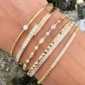 Op de afbeelding: Een verzameling gouden armbanden, elk met een uniek ontwerp. Sommige zijn versierd met diamanten, terwijl andere een getextureerd of parelachtig oppervlak hebben. De armbanden worden op een pols getoond, waarbij hun verschillende stijlen en glans worden getoond.