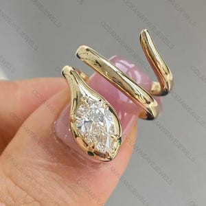 May include: A gold-colored snake ring with a pear-shaped, clear gemstone as the head. The ring wraps around the finger with a coiled design. Small green gemstones accent the head.
