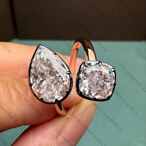 May include: Two rings with large, clear gemstones. One ring features a pear-shaped stone, the other a square-cut stone. Both stones are set in a dark metal with a rose gold band. The rings are held by a hand.